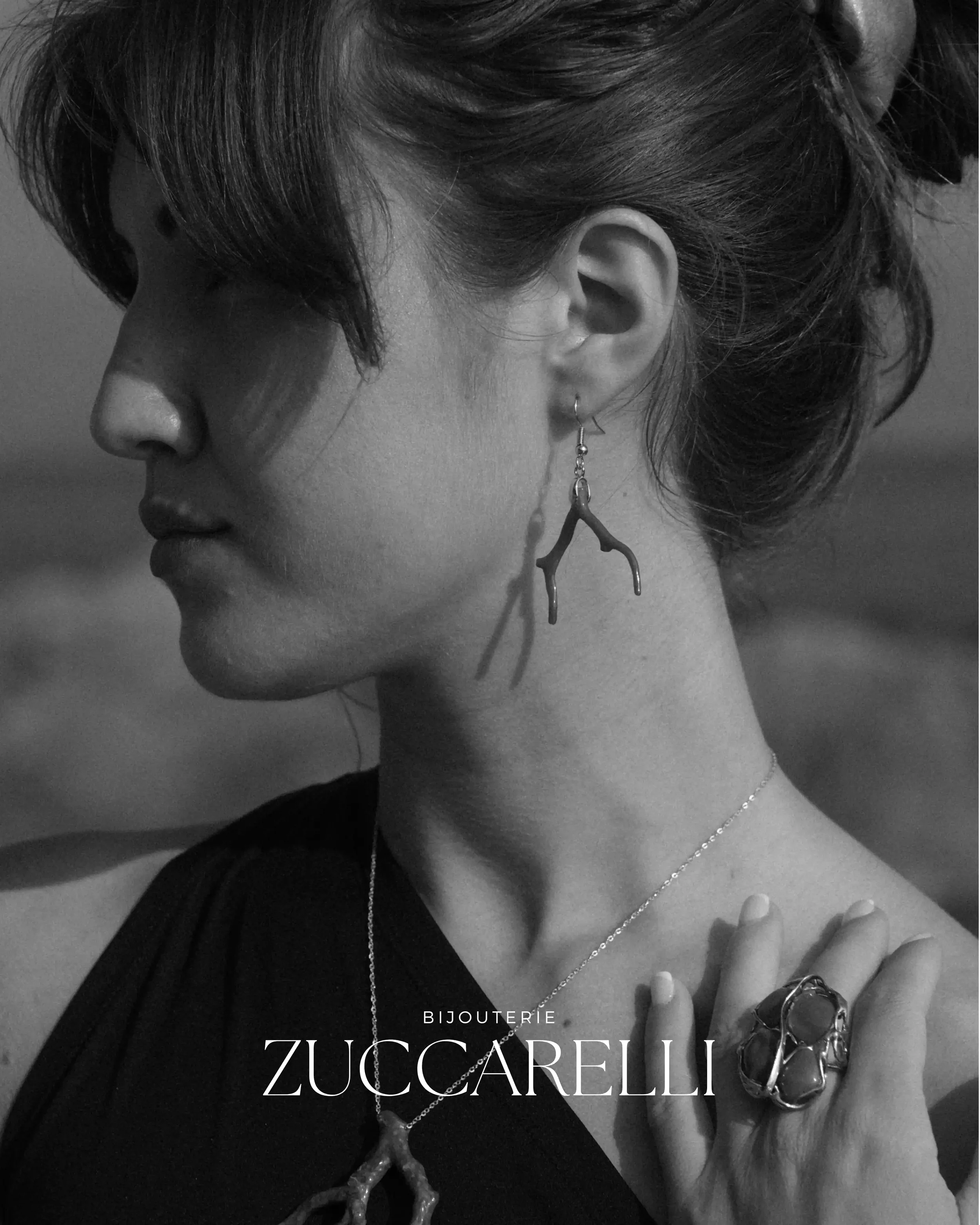 Une femme avec une coiffure courte, portant un bijou avec un pendentif en forme d'arbre et une boucle d'oreille en forme de croissant. La photo est en noir et blanc avec un texte en surimpression indiquant : 'Bijouterie Zucarelli'.