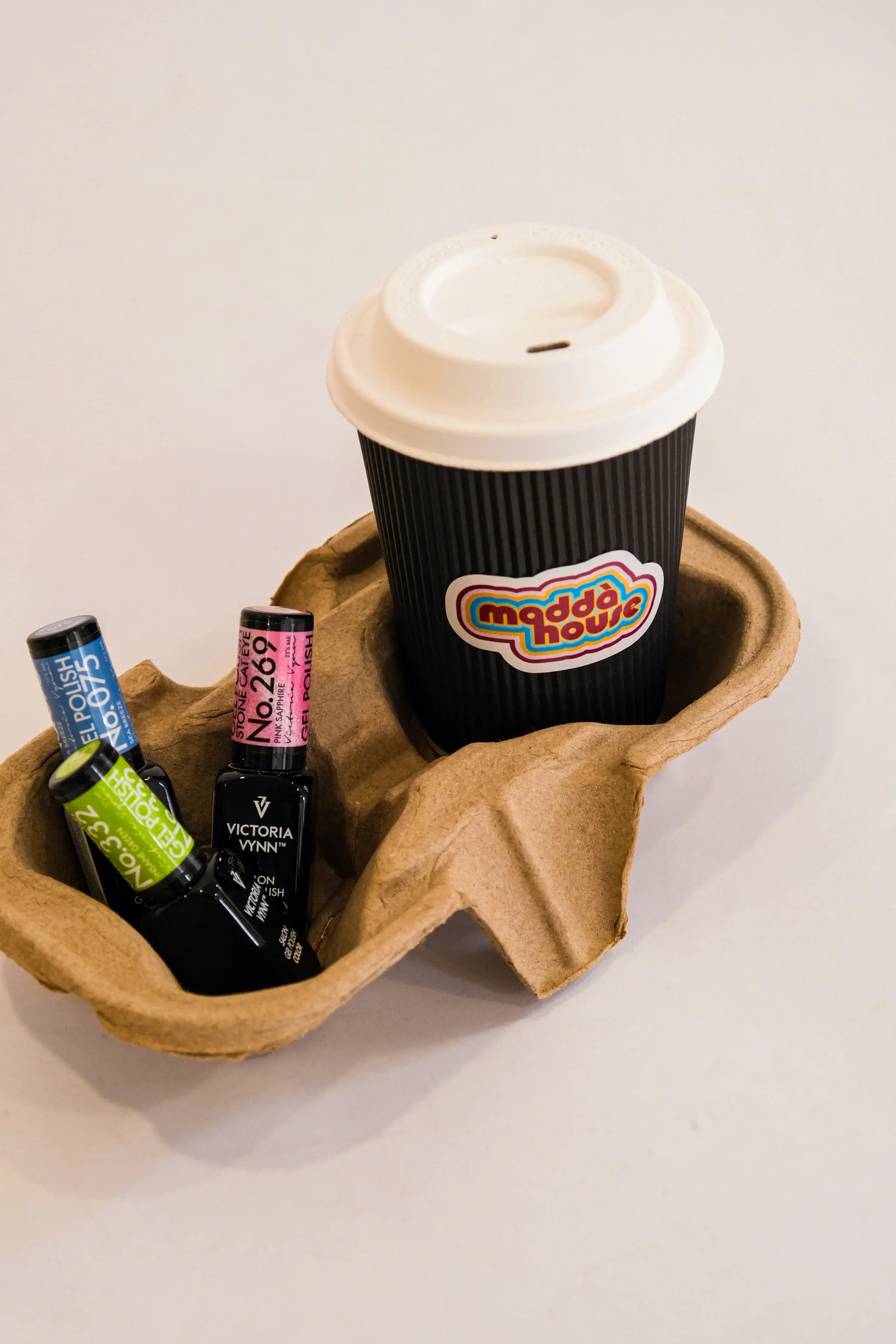 Une boîte en carton contenant plusieurs vernis à ongles avec des couleurs variées et un gobelet en papier noir avec un autocollant "madda house".