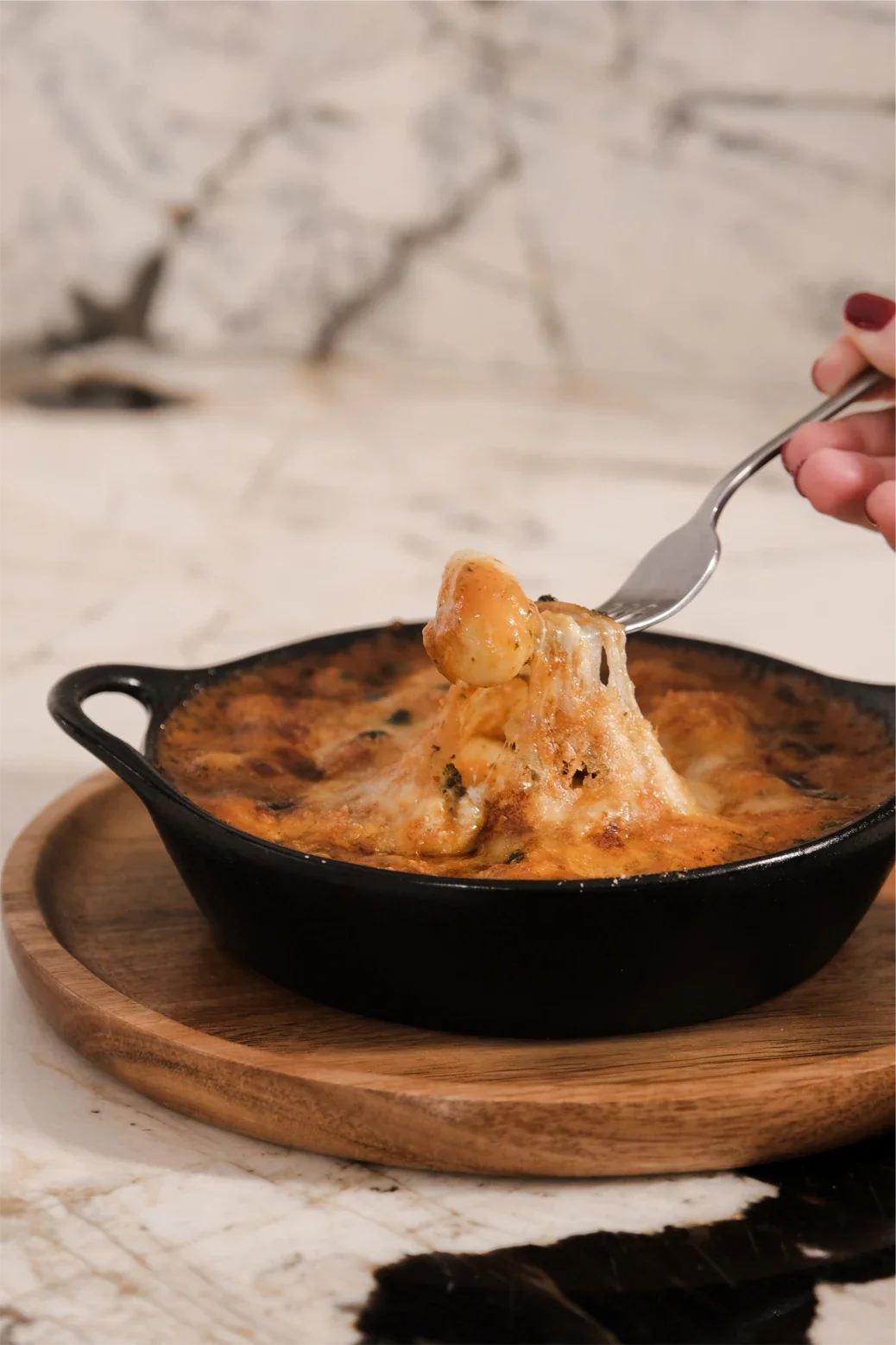 Gratin de pâtes à la sauce tomate dans un plat en fonte noire, posé sur une planche en bois, avec une cuillère contenant une portion de fromage fondu.
