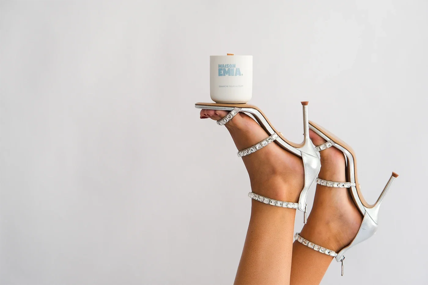 Une main portant une paire de chaussures à talons blancs ornées de strass, avec un pot de crème de visage posé sur la plante du pied, contre un fond neutre.