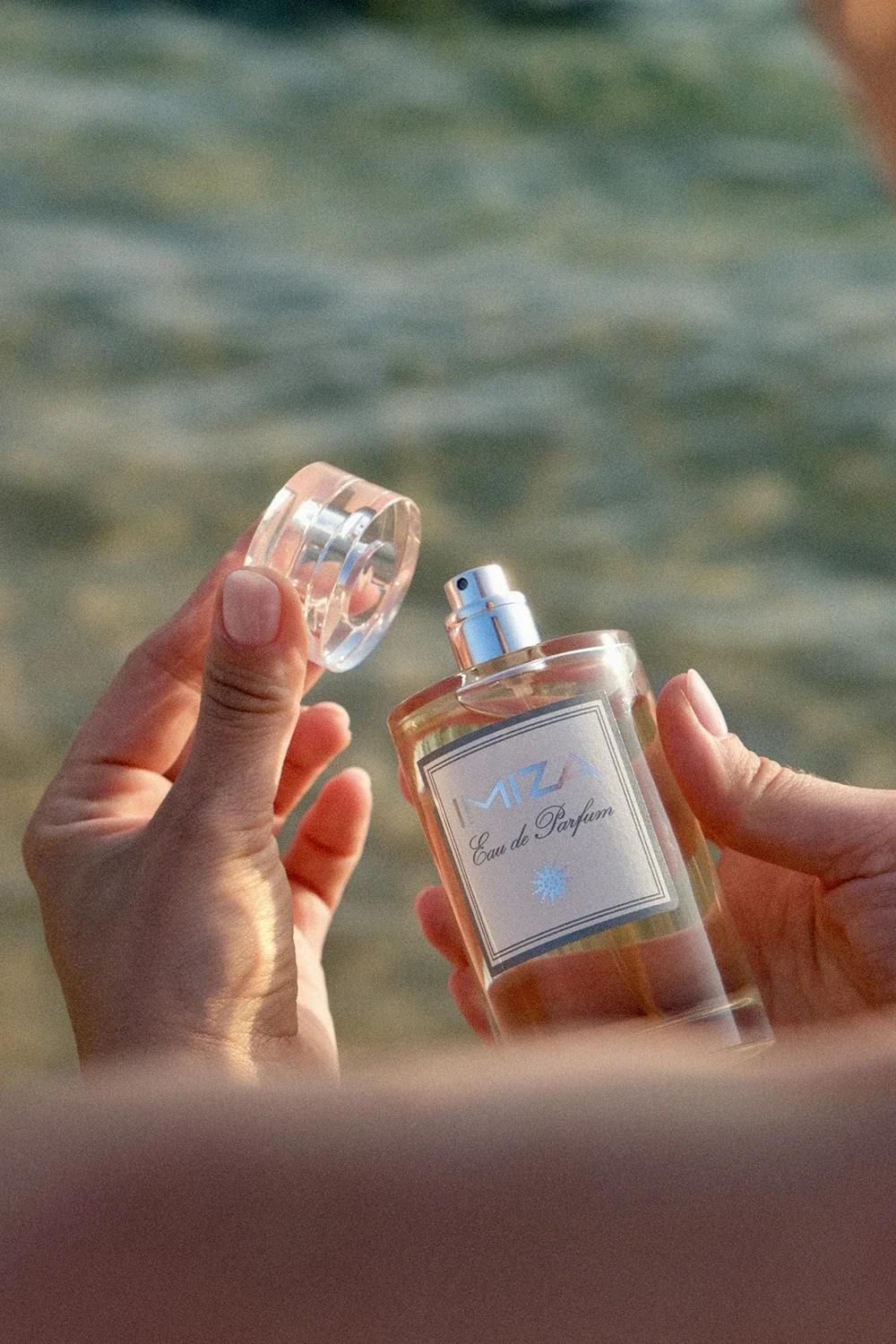 Une personne tient un flacon de parfum ouvert, intitulé "MIZ Eau de Parfum", devant une mer ou un plan d'eau, avec une étiquette blanche et un capuchon transparent