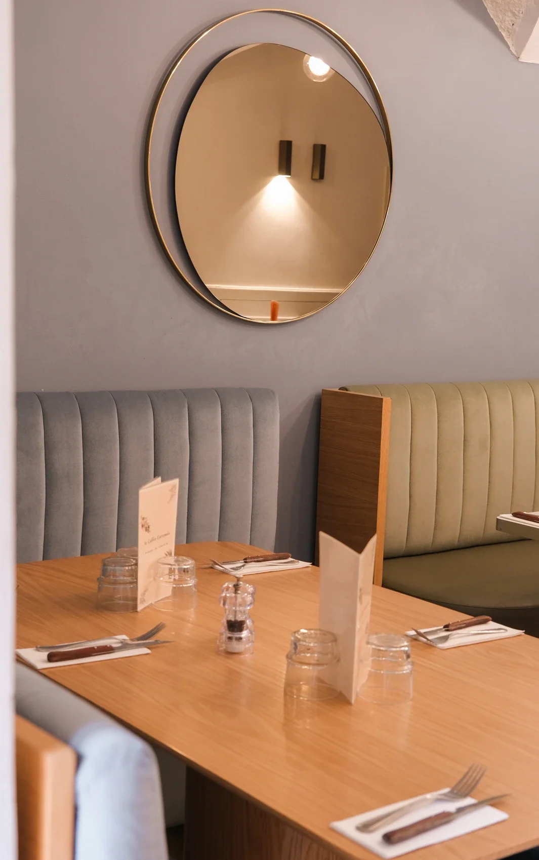 Intérieur d'un restaurant avec des tables en bois, des assiettes, des verres, des couverts en position de service, des menus et des banquettes en tissu gris et vert. Un grand miroir rond avec une bordure dorée est accroché au mur, reflétant un éclair