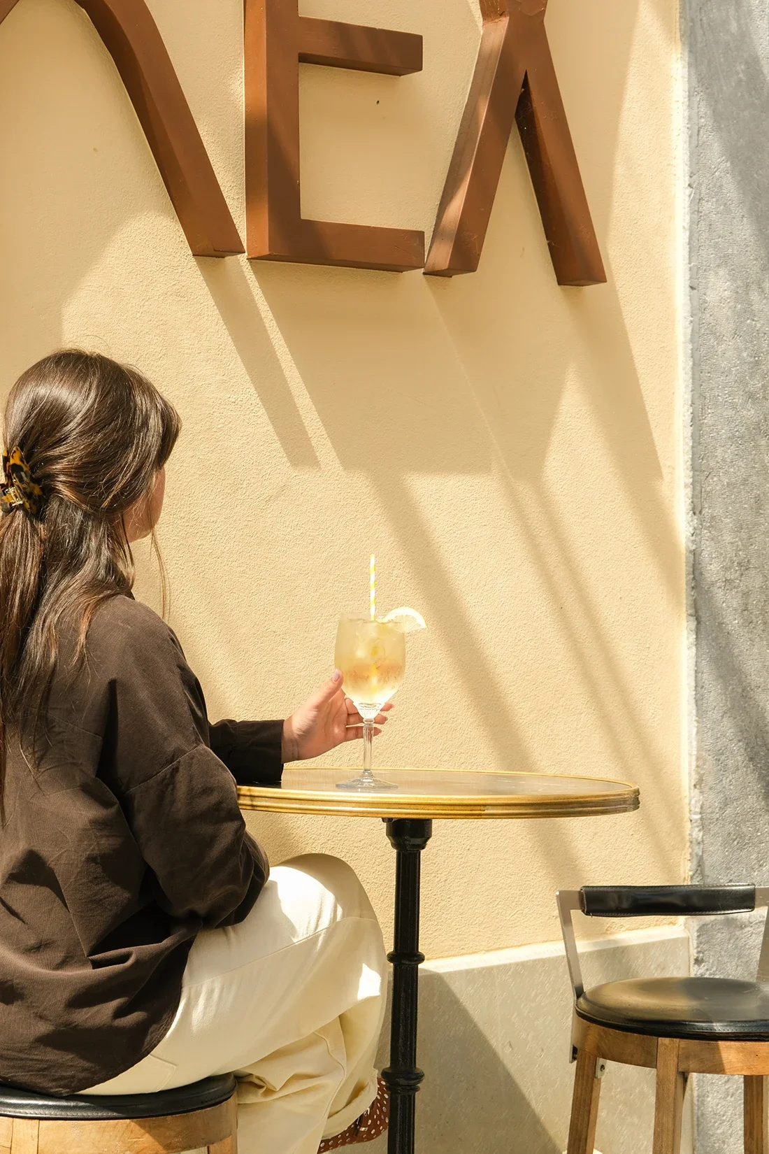 Une femme assise à une table dans un café, tenant un verre avec une boisson glacée, une tranche de citron et une paille. La scène est intérieure avec un mur beige et une décoration en bois.