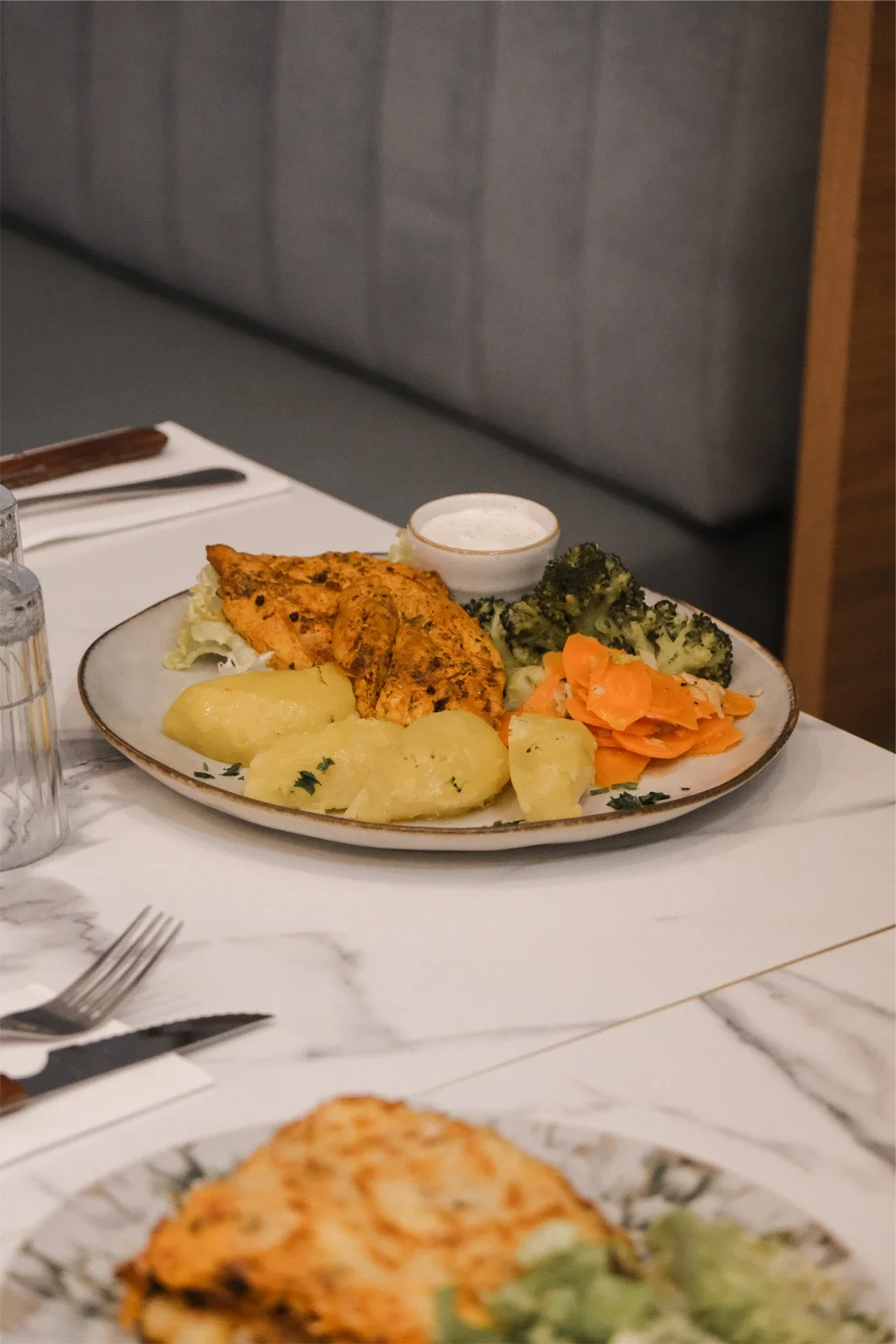 Une assiette avec poisson épicé, pommes de terre, brocolis, carottes râpées, et une sauce à côté, sur une table de restaurant.