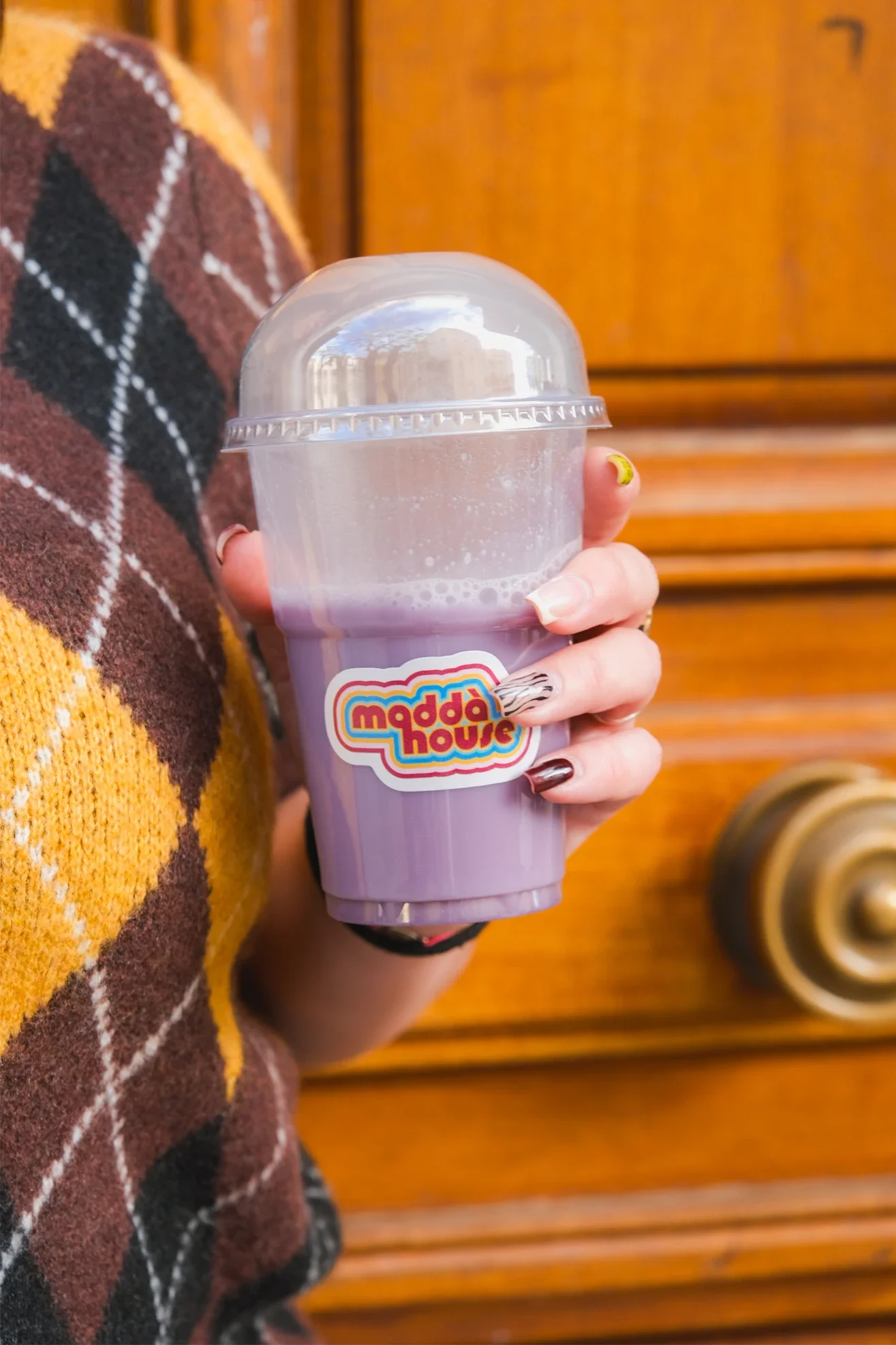 Une personne tient un grand verre de boisson violette avec une étiquette 'madda house'. La personne porte une veste en laine à motif écossais et a des ongles peints. La photo est prise devant une porte en bois.