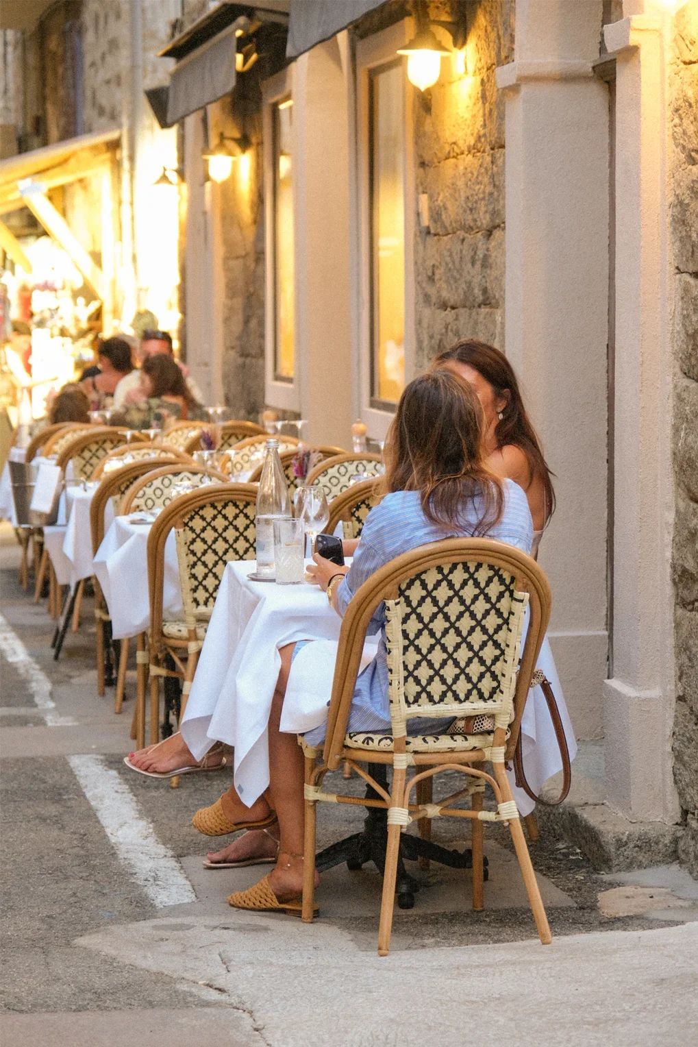 Deux femmes discutent à une table d'un restaurant en plein air, éclairé par des lampes jaunes, avec des chaises en rotin et une table dressée avec des verres et une bouteille d'eau, dans une rue pavée lors de la tombée de la nuit.