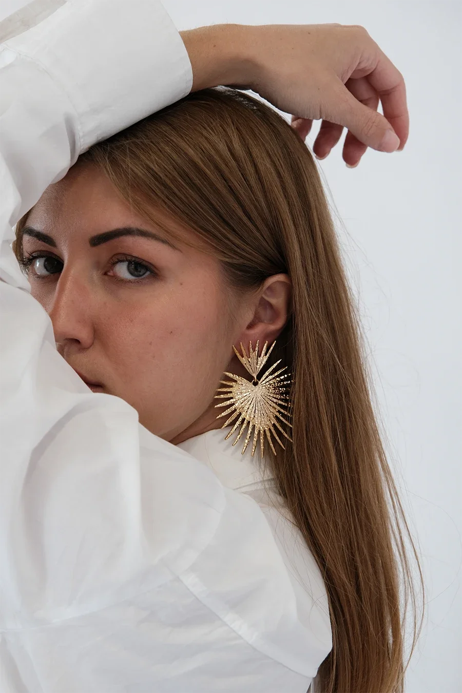 Femme avec boucle d'oreille or grande et vêtements blancs, pose artistique avec main sur la tête, fond blanc.