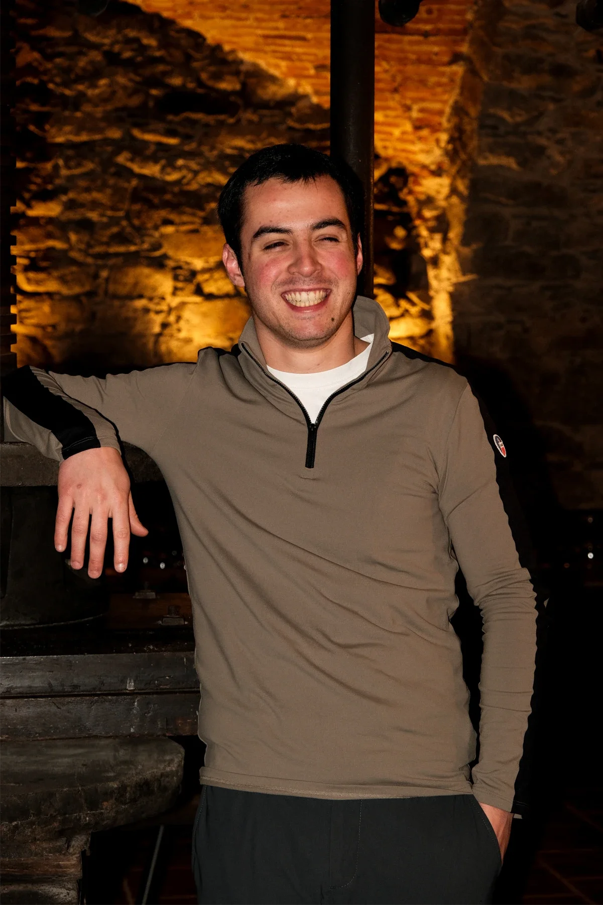 A smiling young man with dark hair, wearing a beige and black long-sleeved athletic shirt, posing indoors in front of a stone wall with warm lighting.
