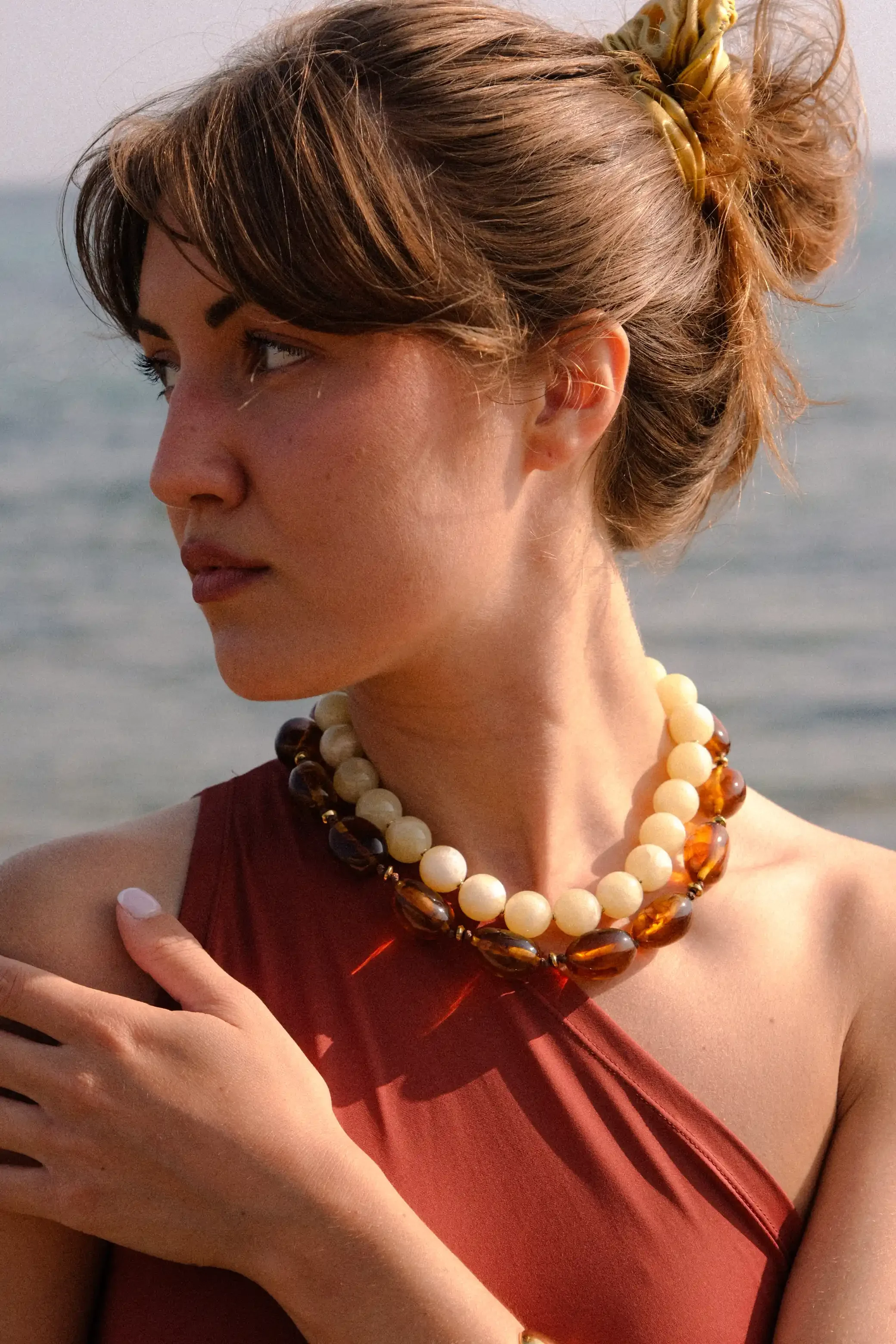 Une femme aux cheveux bruns bouclés, portant un chignon avec un ruban vert, porte un collier de perles beige et marron, et une robe rouge foncé, capturée lors d'un coucher de soleil près de la mer.