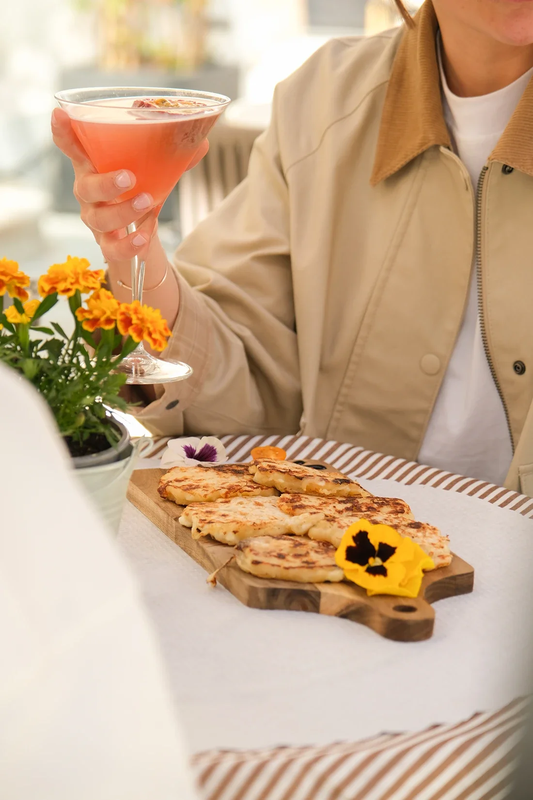 Une personne tient un verre de cocktail rose à la main, assise à une table avec des muffins chauds sur une planche en bois, décorée de fleurs colorées. L'ambiance est lumineuse et chaleureuse.