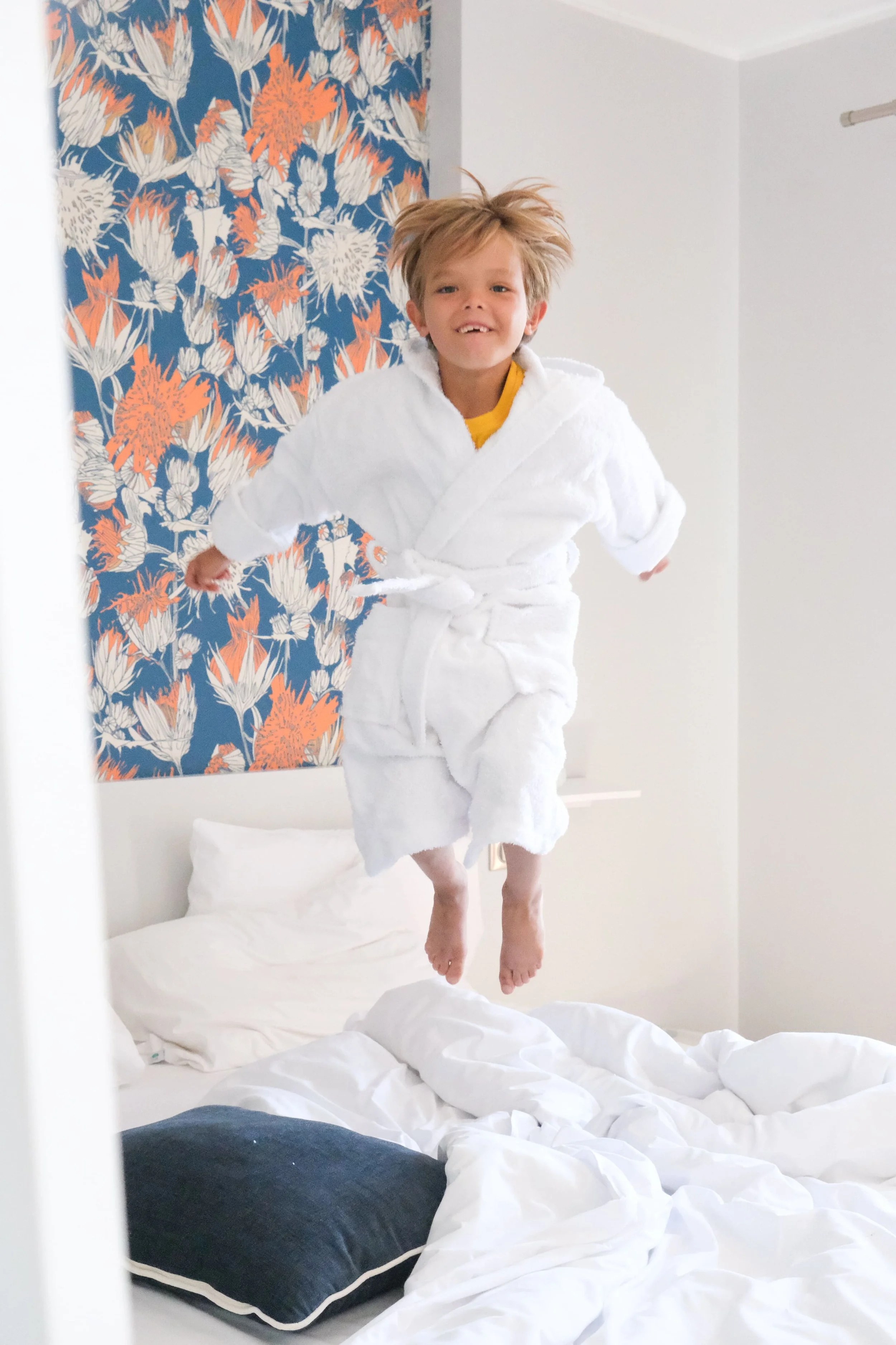 Un enfant qui saute sur un lit d'hôtel. L'enfant porte un peignoir blanc. Sur le lit, il y a une couette blanche, et des coussins.