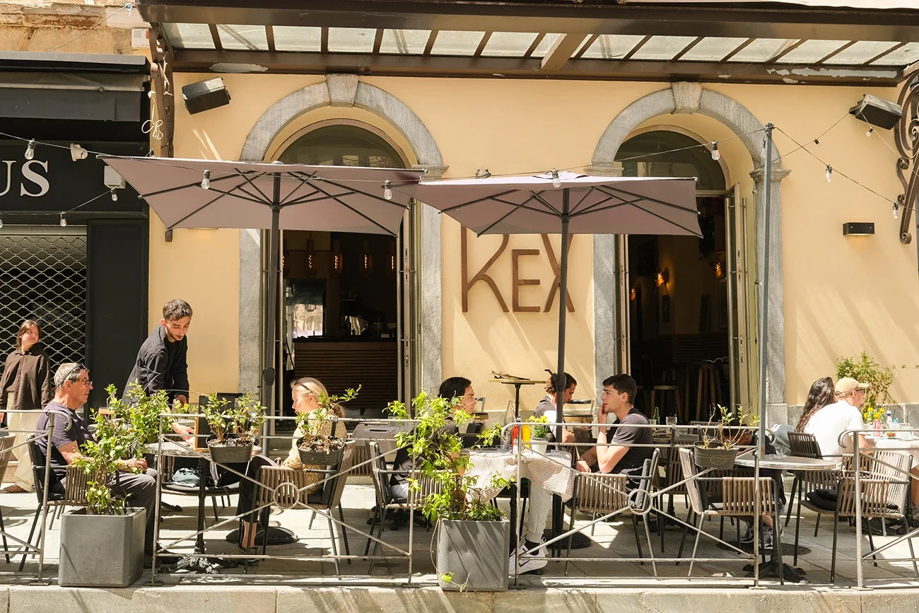 Terrasse de café ou restaurant avec des tables et des chaises, parasols, et plusieurs clients dégustant des repas ou des boissons, devant un bâtiment avec deux arches et la lettre 'KEX' sur le mur.