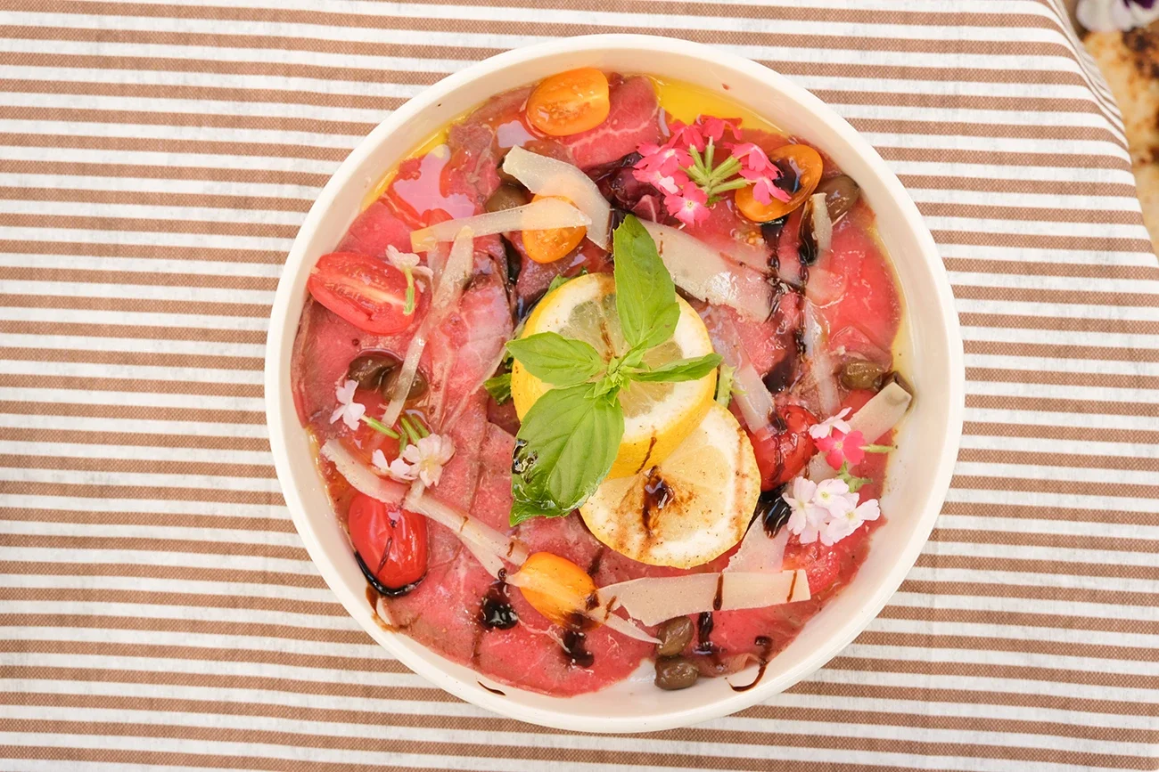 Salade de carpaccio de bœuf avec des tomates, olives, citron, basilic, fleurs comestibles, et assaisonnements divers, vue de dessus sur une serviette à rayures.