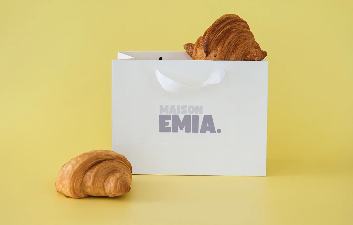 Une boîte blanche de la marque Maison Emia avec deux croissants, un dans la boîte et un devant. Fond jaune.