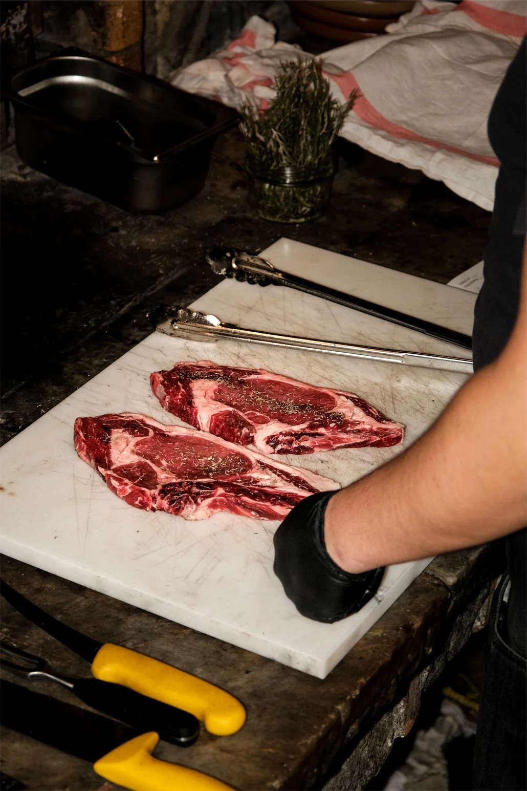 Deux steaks crus sur une planche à découper, assaisonnés avec des herbes. Tagueur noir portant des gants noirs à côté, ustensiles de cuisine en arrière-plan, un moule en verre avec des herbes fraîches et un chiffon blanc avec des rayures rouges sur u