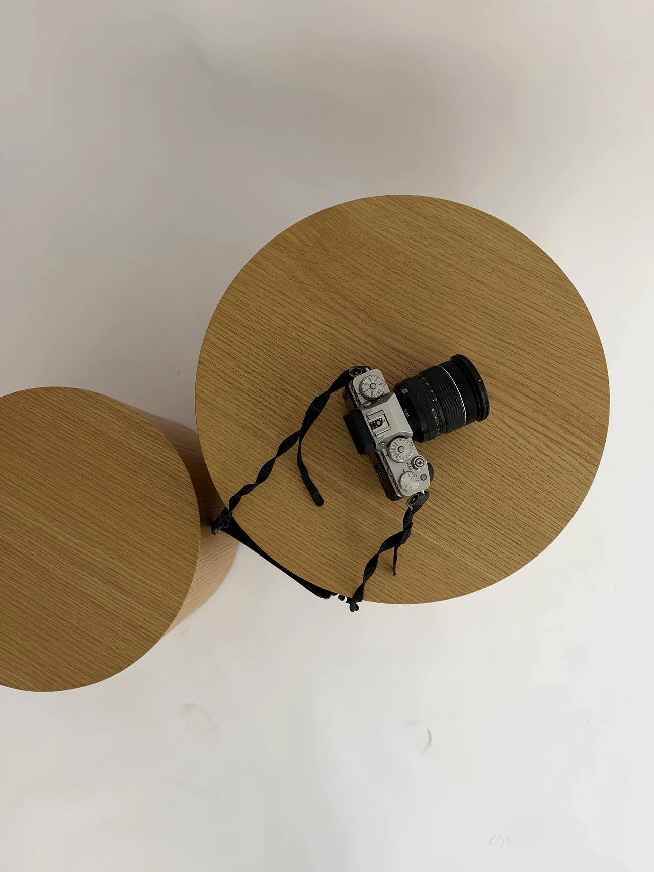 Un appareil photo argenté avec un objectif noir repose sur une table ronde en bois clair. La table est placée sur fond blanc, avec un autre cercle en bois clair à côté.