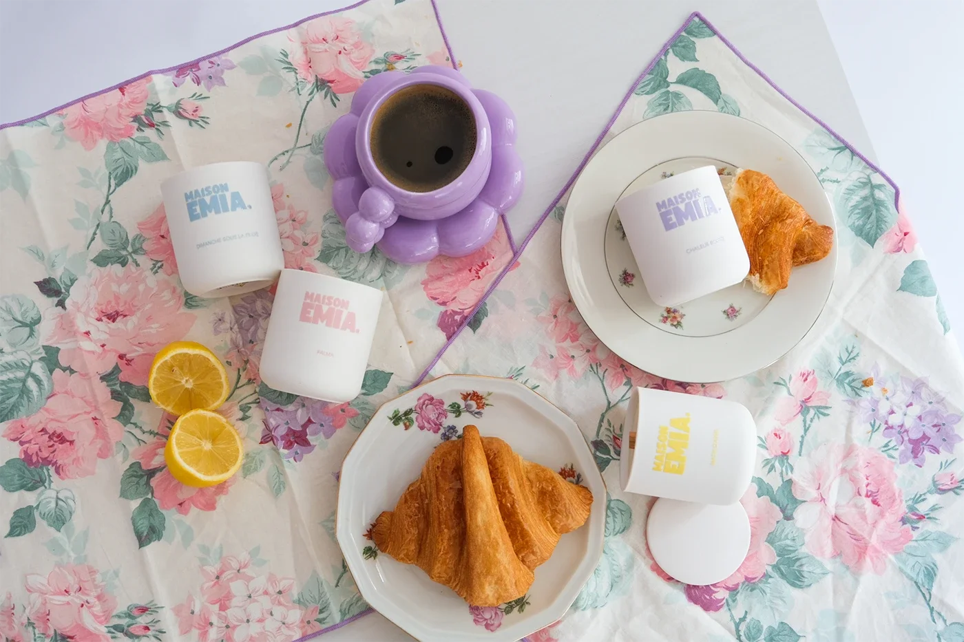 Petit-déjeuner avec deux croissants, une tasse de café, deux tasses avec le logo 'MAISON EMIA', un verre de citron et une tasse violet en forme de bouquet de fleurs, sur une table avec une nappe à fleurs