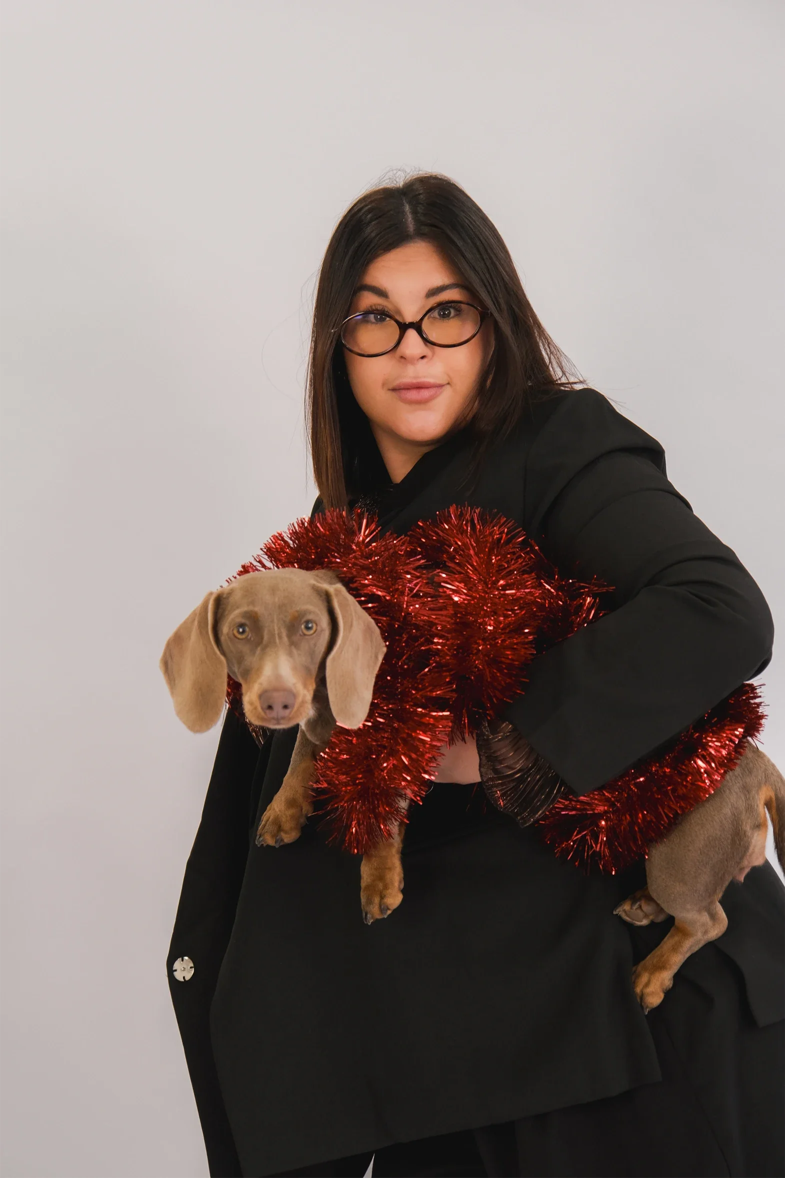 Une femme avec des lunettes tient un chien de race vizsla, tous deux décorés avec une guirlande rouge brillante, en contrebas d'un fond blanc.