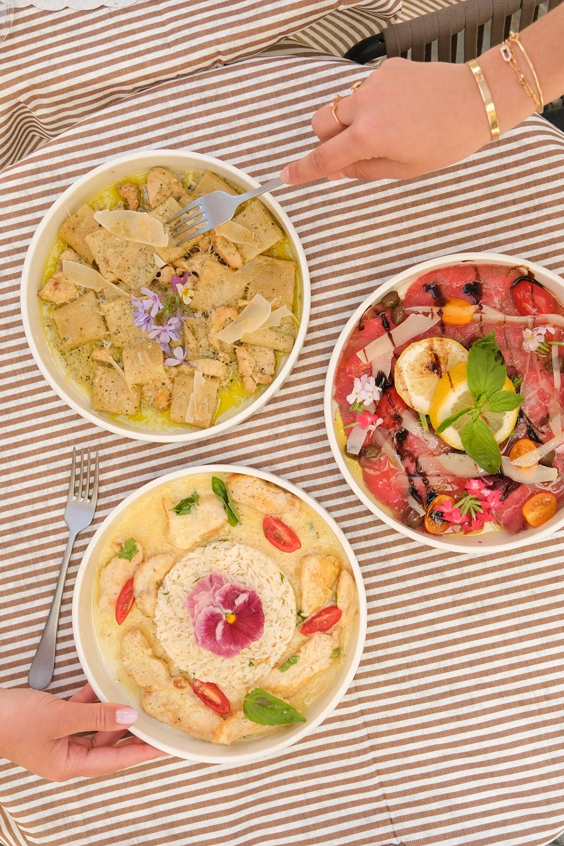Trois plats de nourriture méditerranéenne ou italienne sur une table à rayures brunes et blanches. Un plat semble contenir des pâtes avec des fleurs comestibles, un autre un carpaccio de viande avec des tomates cerises et des herbes, et le troisième 