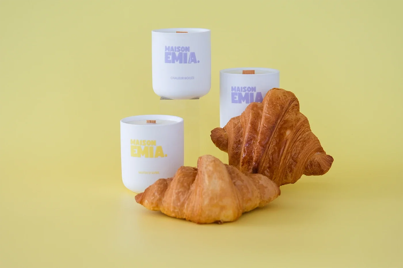 Trois bougies parfumées de la marque Maison Emia dans des contenants blancs, accompagnées de deux croissants.