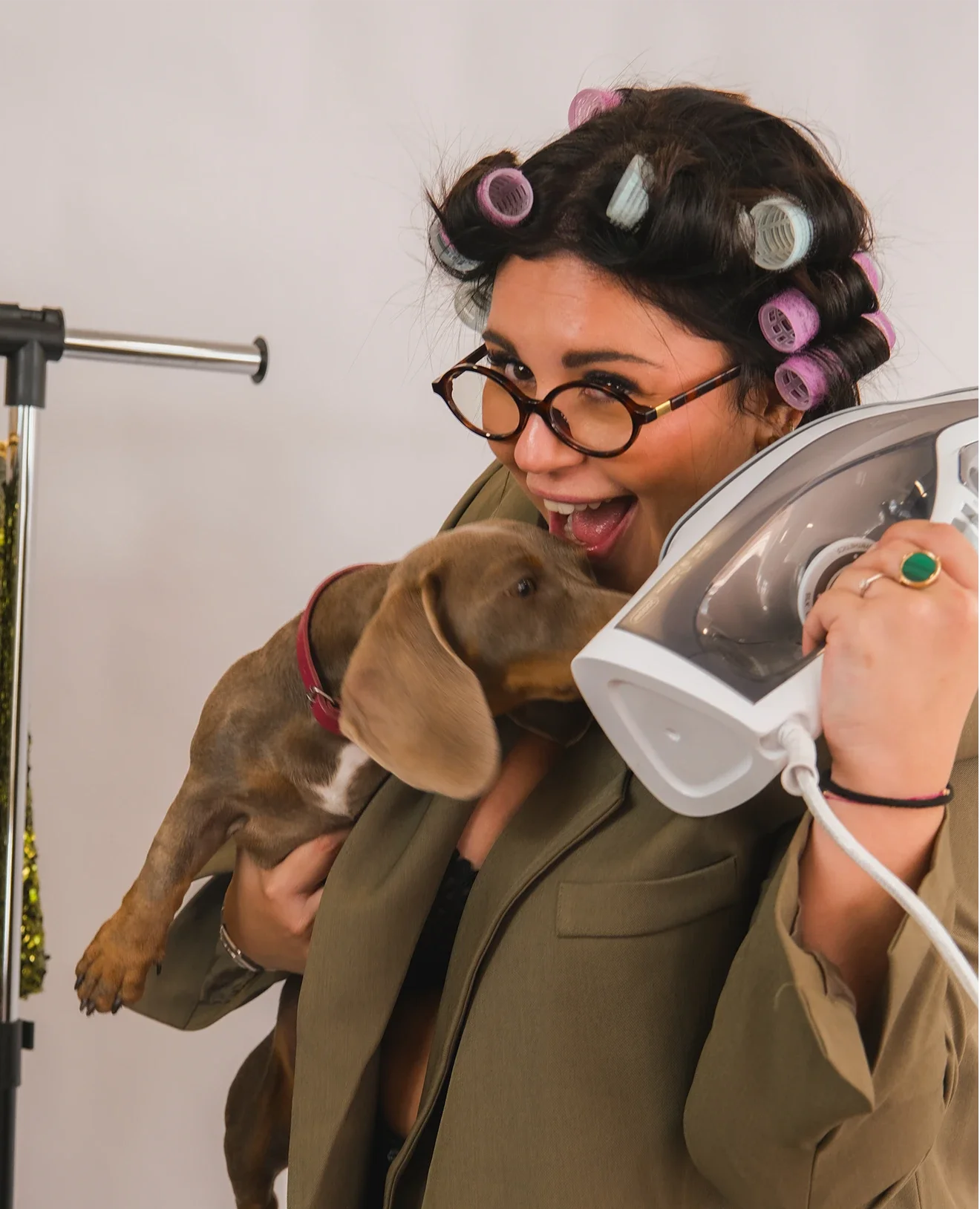 Une femme avec des rouleaux dans les cheveux, portant des lunettes, tient un fer à repasser et un chiot coloré, dans un intérieur 