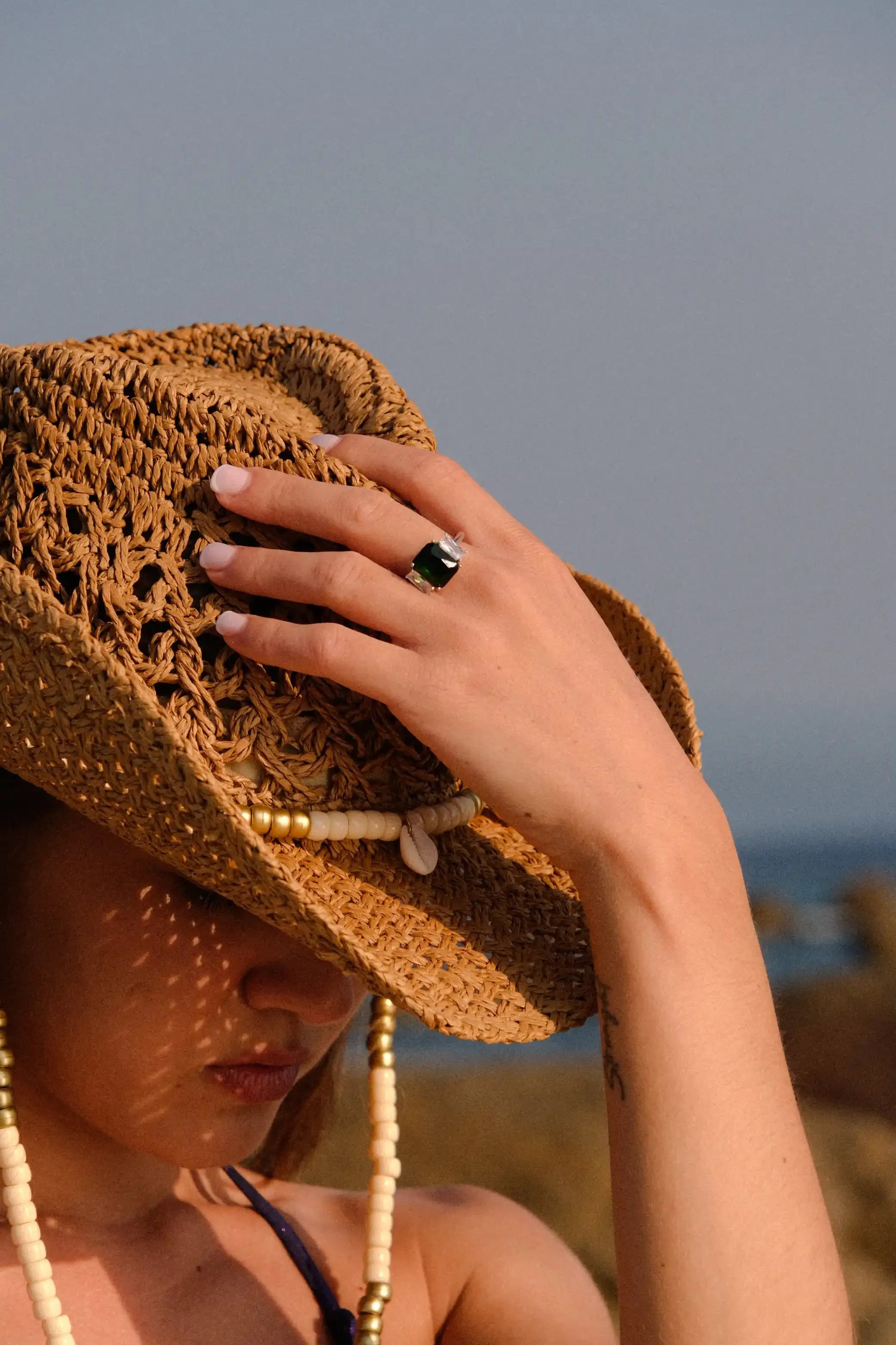 Une femme portant un chapeau de paille tressé, avec des perles blanches, un collier noir et une robe noire, tenant le chapeau devant son visage, avec une peau bronzée et des tatouages, en extérieur