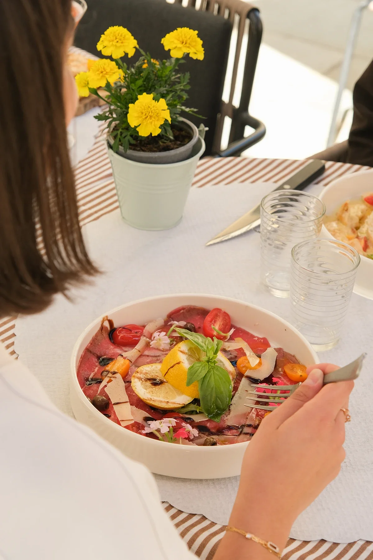 Une personne mange une salade froide avec des légumes, des œufs et des herbes sur une table avec un pot de fleurs jaunes.