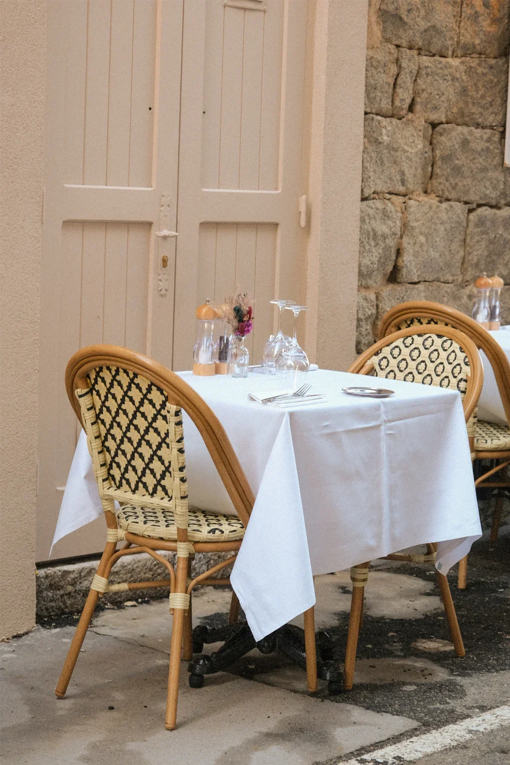 Table de restaurant avec vaisselle, verres, couverts et un vase avec des fleurs, située à l'extérieur près d'un mur en pierre et d'une porte en bois blanc.