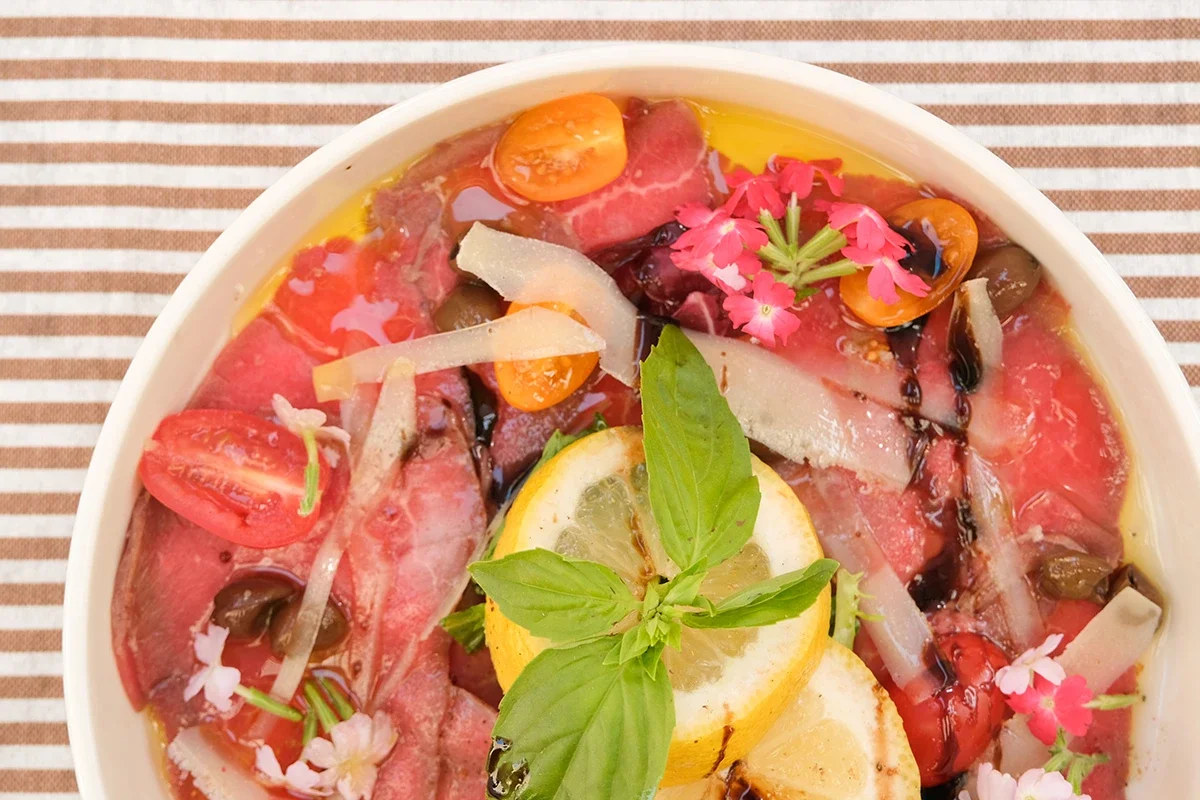 Carpaccio de viande avec des tranches de citron, des tomates, des herbes, et des fleurs comestibles.
