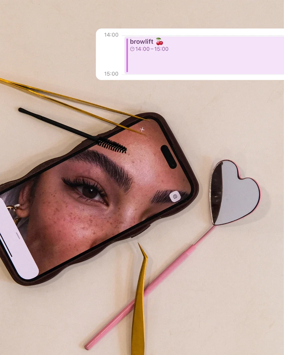 Un téléphone affichant un œil d'une personne avec des sourcils très épais, avec deux outils de beauté pour les sourcils, un miroir en forme de cœur, et une conversation de planification de séance de lifting des sourcils 'brow lift'.