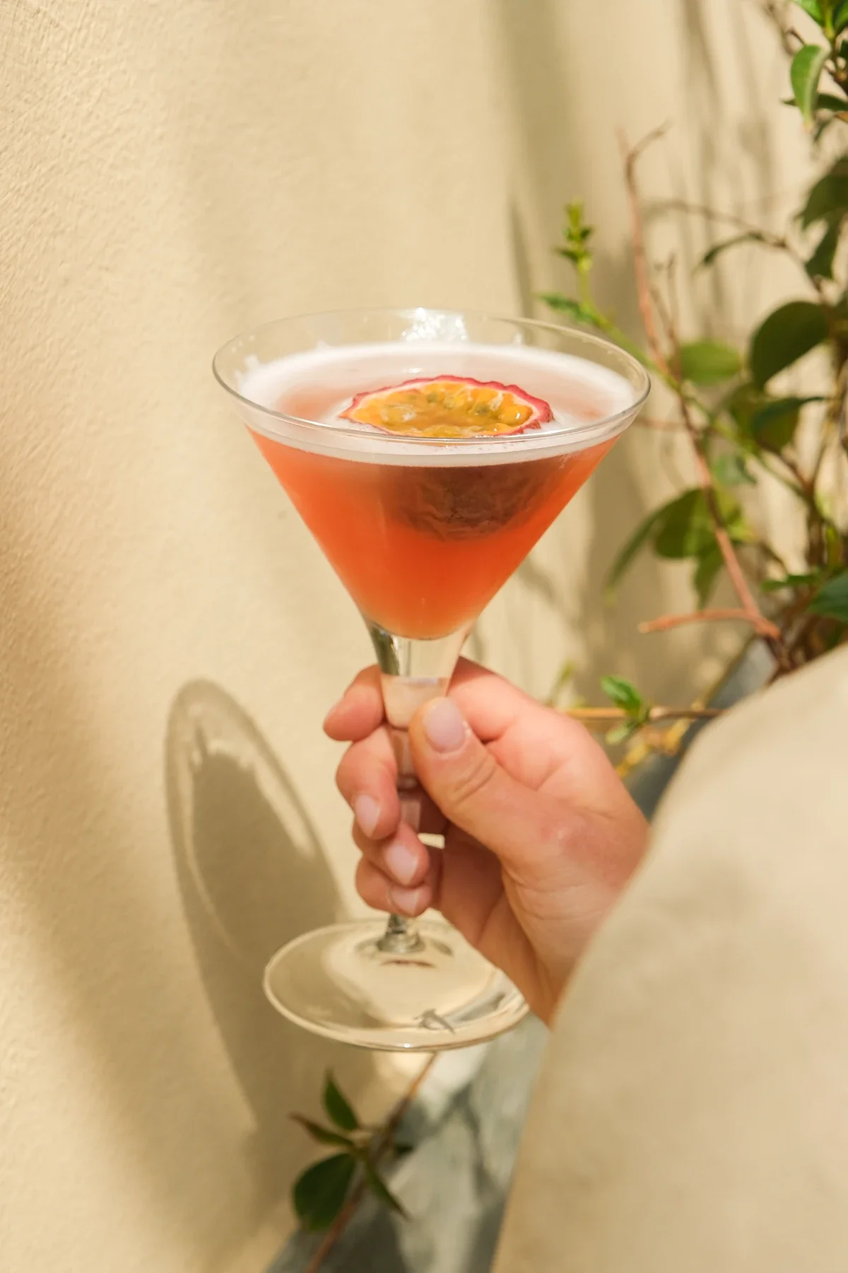 Un verre à cocktail en main avec une boisson rose, garnie d'une tranche de fruit passiflore flottant dessus.