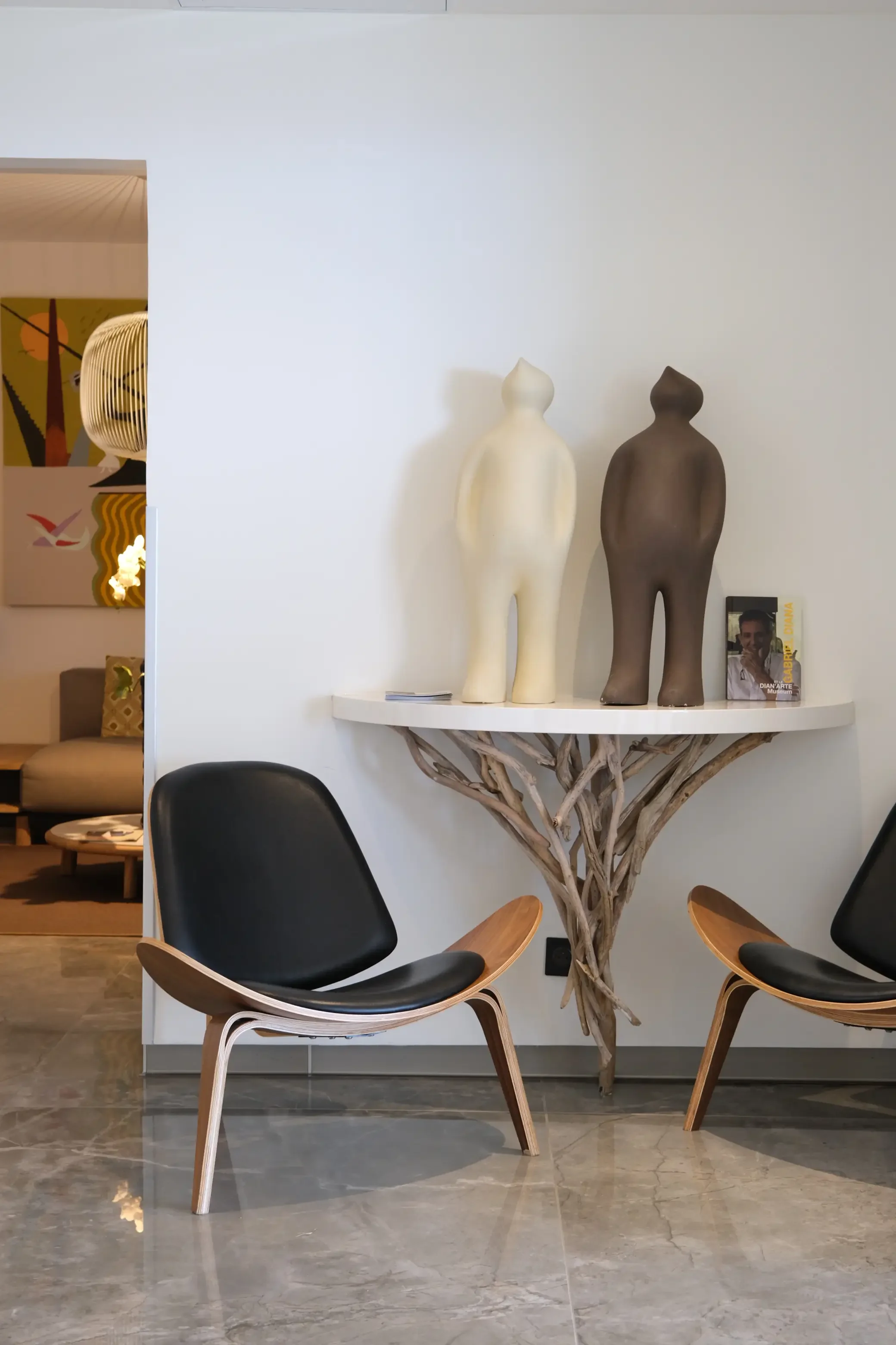 Deux sculptures de silhouettes humaines, l'une blanche et l'autre brune, posées sur une étagère contre un mur blanc, avec un livre posé à côté de la sculpture brune, dans un intérieur moderne avec fauteuils en bois et cuir.