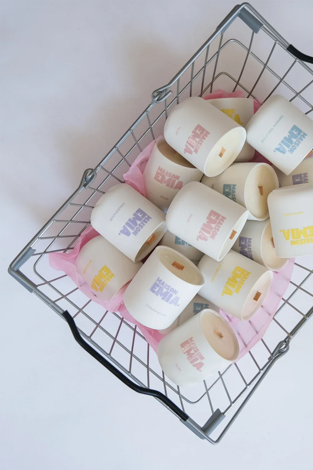 Un panier en métal contenant plusieurs tasses blanches avec le logo coloré "VUE MAISON" en pastel dans différentes couleurs, posé sur un fond blanc avec du papier rose en dessous.