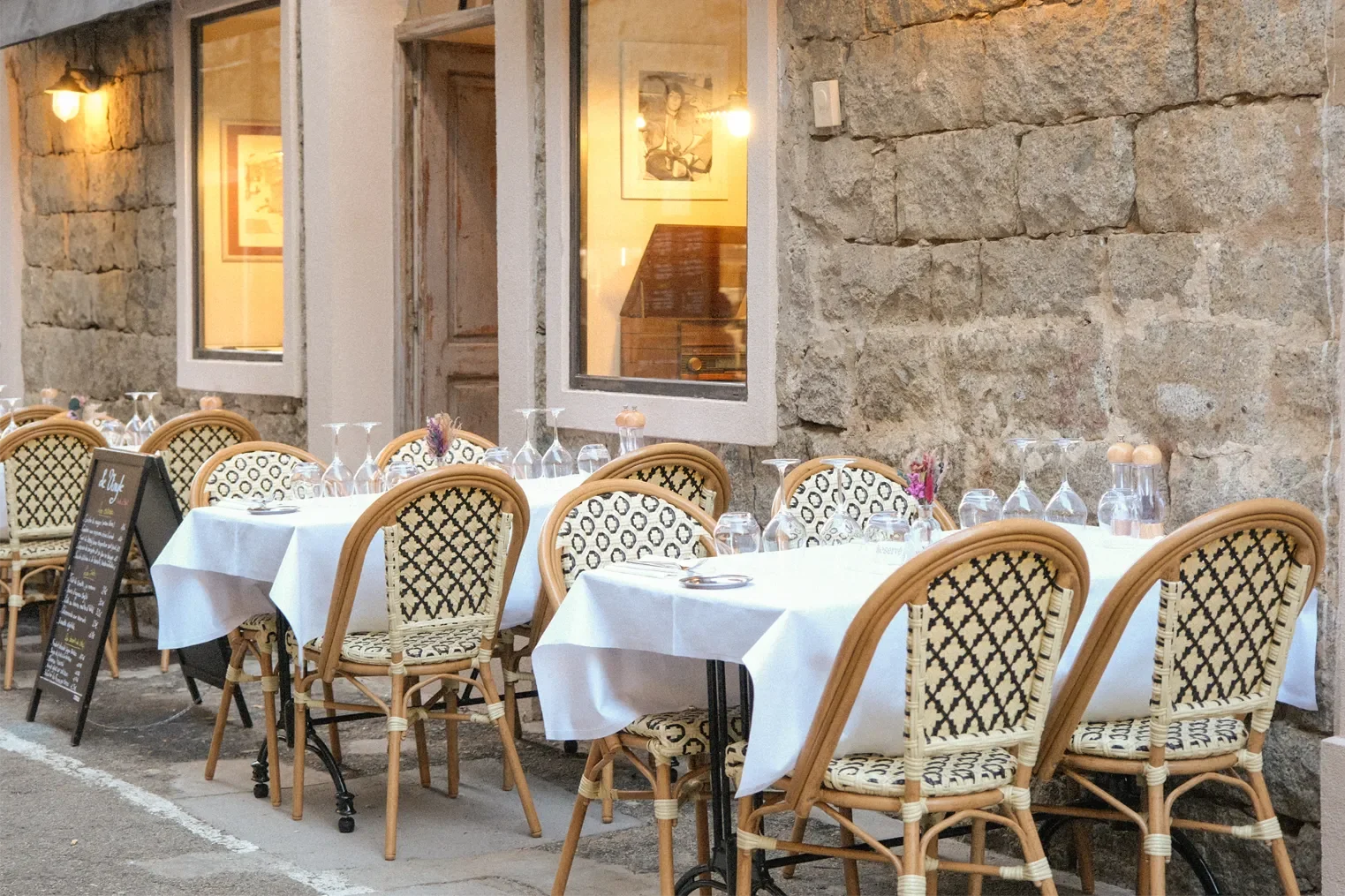 Terrasse de restaurant avec tables habillées de nappes blanches, chaises en rotin avec coussins à motifs, et menu au tableau noir. Mur en pierre avec fenêtres donnant sur un intérieur lumineux.