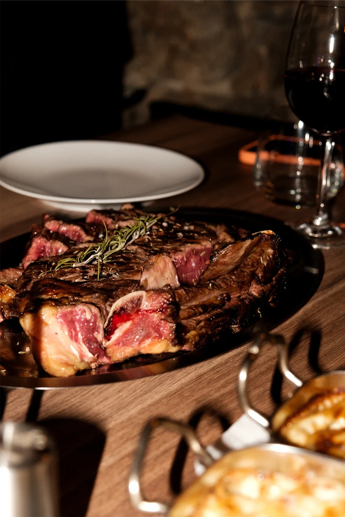 Une assiette de viande grillée avec des herbes, une assiette blanche vide, un verre de vin rouge, et un fond de mur en pierre dans une ambiance chaleureuse.