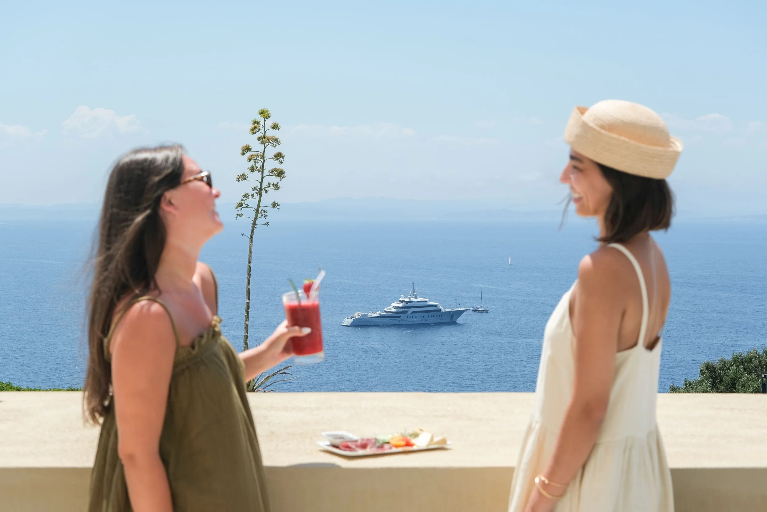 Deux personnes qui rient, avec une planche repas au milieu. Une personne porte une robe kaki, une paire de lunettes et tient une boisson dans sa main. L'autre personne porte une robe beige et un chapeau. En fond la mer et un bateau.