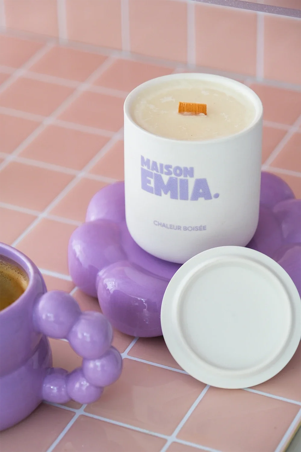 Bougie parfumée blanche avec inscription en violet, posée sur un support violet en forme de chaîne de perles, dans un décor de carreaux rose pâle.