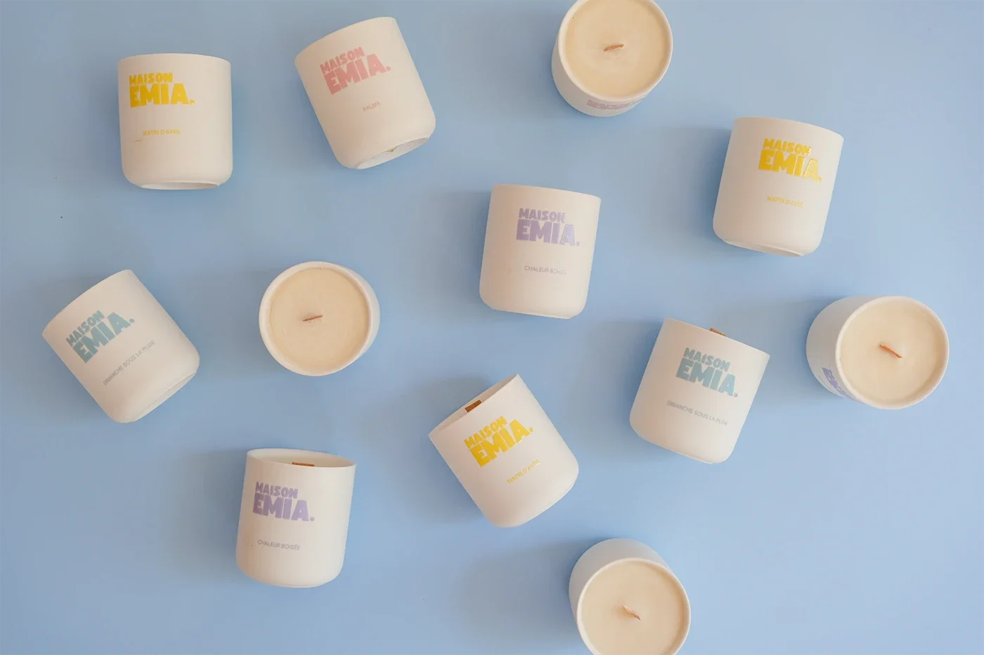 Bougies parfumées dans des pots blancs avec étiquettes colorées, disposées sur une surface bleu pastel.
