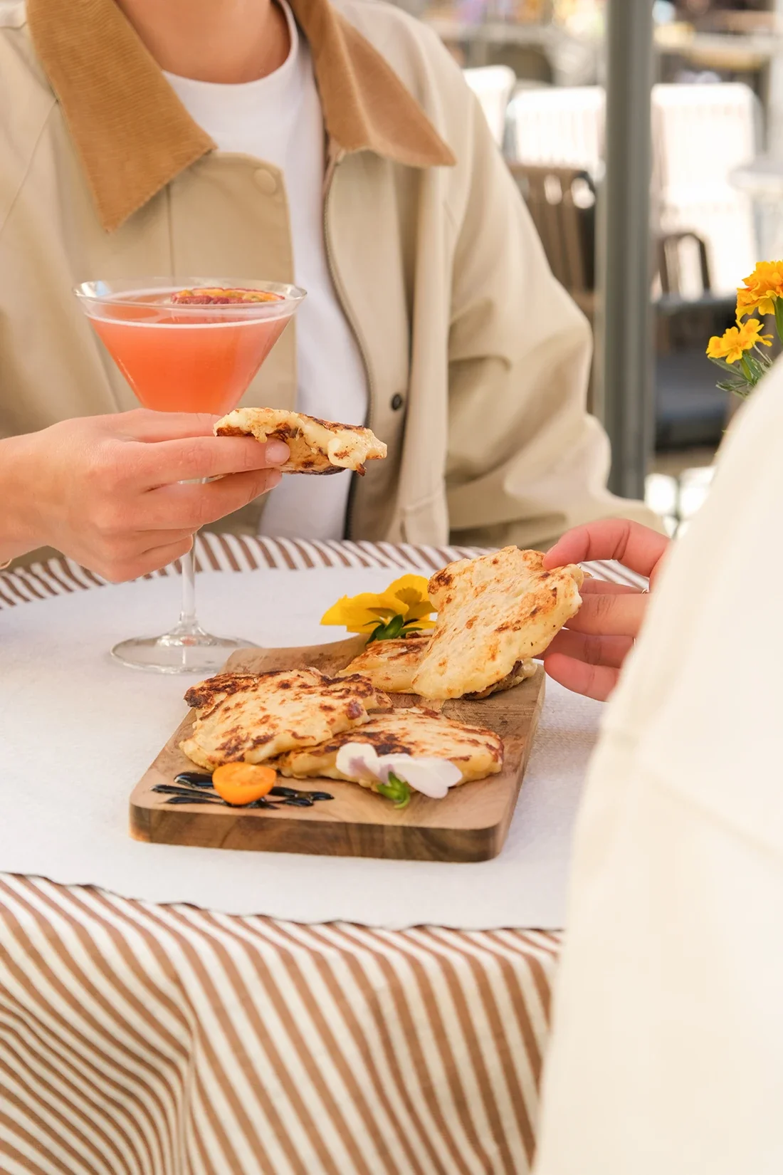 Une personne tient un verre à cocktail rose avec une tranche d'orange, et une autre personne tient une part de pizza. Sur la table, un plateau en bois avec plusieurs parts de pizza, garnies de fromage fondu, de tomates cerises et de fleurs.