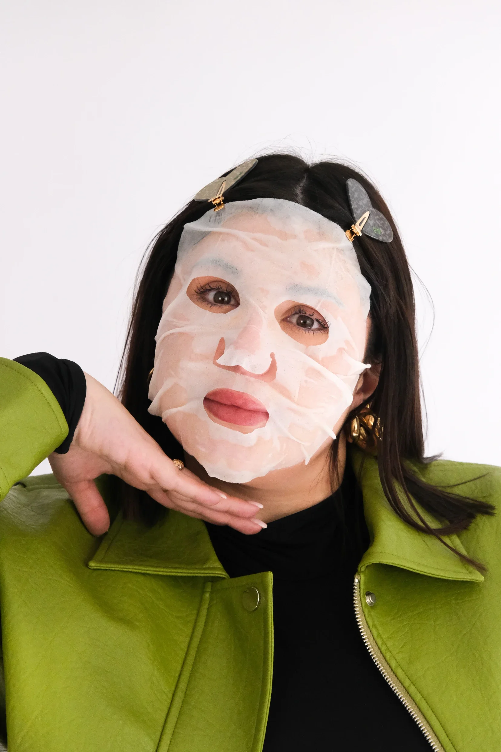 Femme portant un masque facial blanc avec des papillons gris clips dans ses cheveux noirs, portant un blouson vert vif.