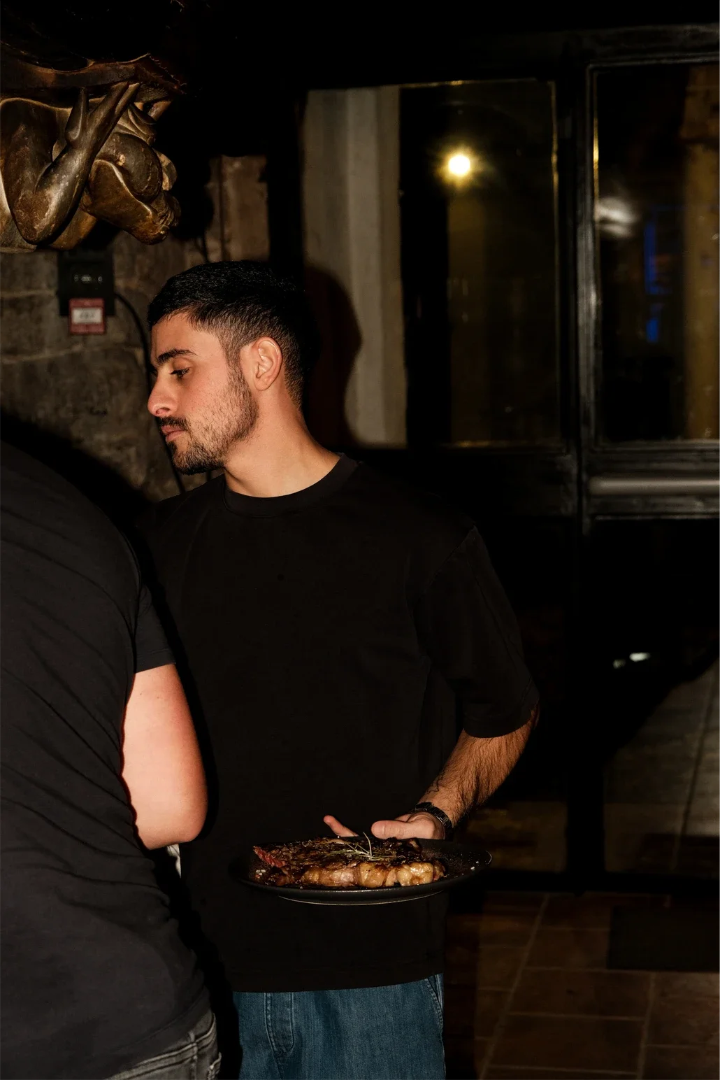 Jeune homme en tenue noire tenant une assiette de viande, dans un restaurant ou un bar, décor en briques et sculpture murale