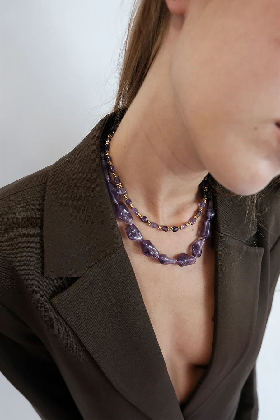 Un gros plan du cou et du décolleté d'une femme portant deux colliers en perles violettes avec des détails dorés, habillée en veste marron foncé.