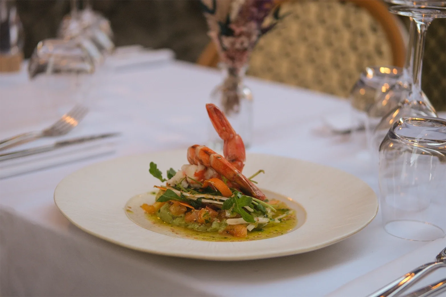 Une assiette de fruits de mer avec une crevette, entourée de légumes verts et de sauce, sur une nappe blanche dans un restaurant élégant.