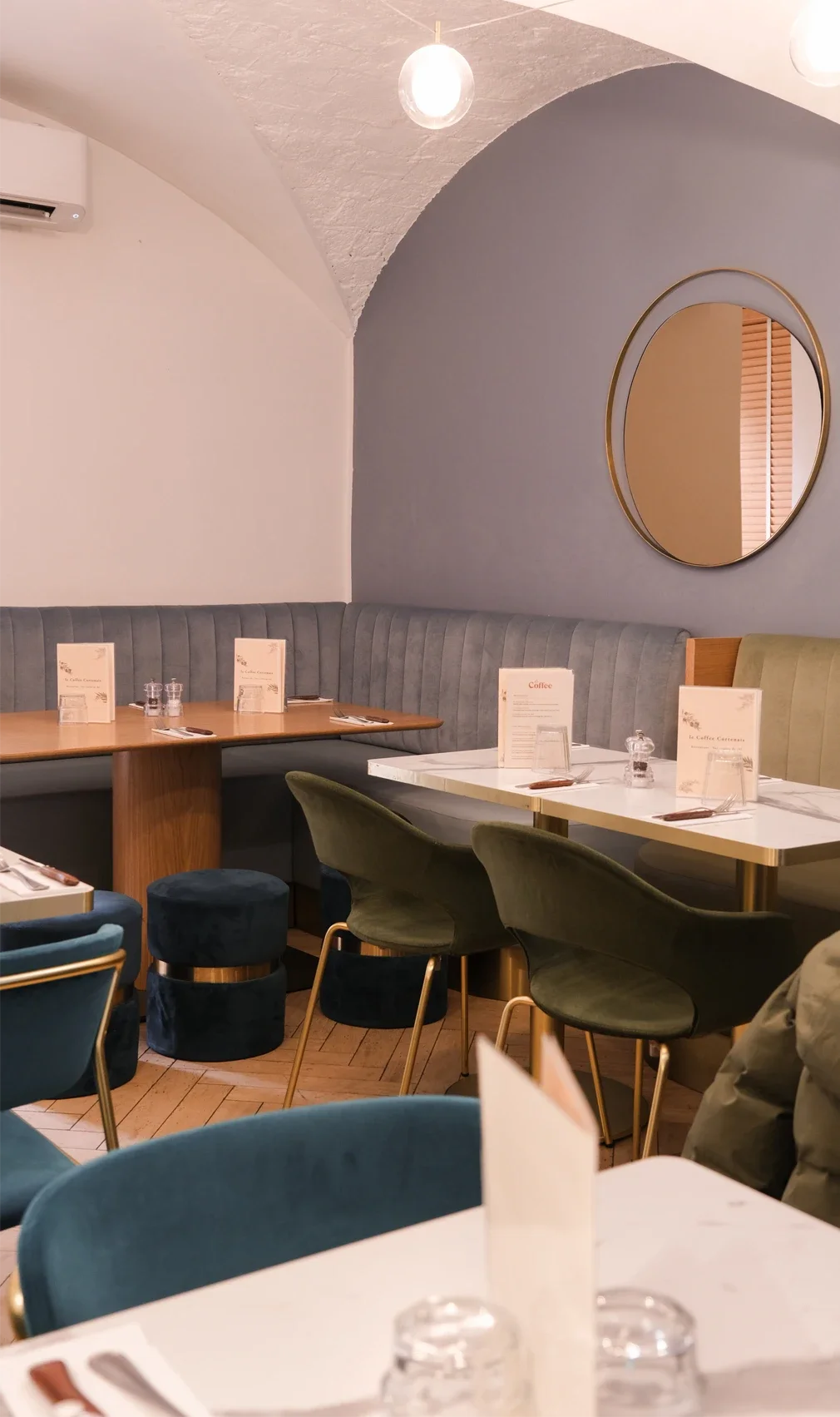 Une salle à manger de restaurant avec des tables, des chaises vertes et bleues, un grand miroir rond, un panneau lumineux blanc, et une décoration élégante.