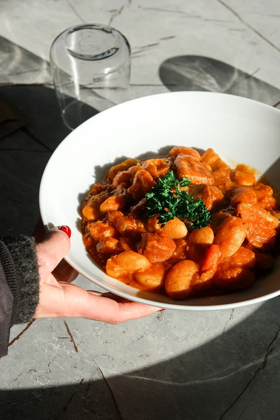 Un plat de ragoût de viande et de haricots, garnie de persil, tenu par une personne avec des ongles rouges, sur une surface en pierre avec un verre à vin vide en arrière-plan.