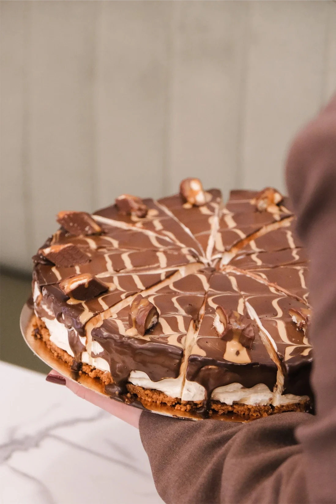 Gâteau au chocolat décoré de morceaux de chocolat et de filets de chocolat blanc, présenté sur un plat blanc, tenu par une personne en vêtement marron