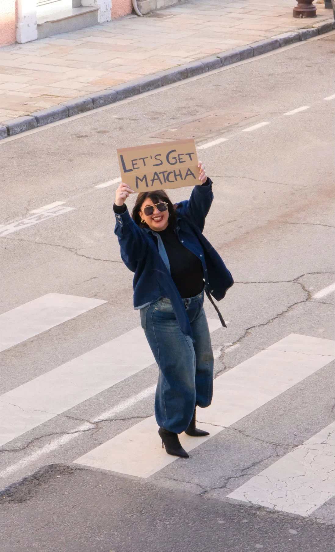 Une femme souriante portant des lunettes de soleil, un jean et un manteau bleu, tient une pancarte avec l'inscription "Let's Get Matcha" tout en marchant sur un passage piéton.