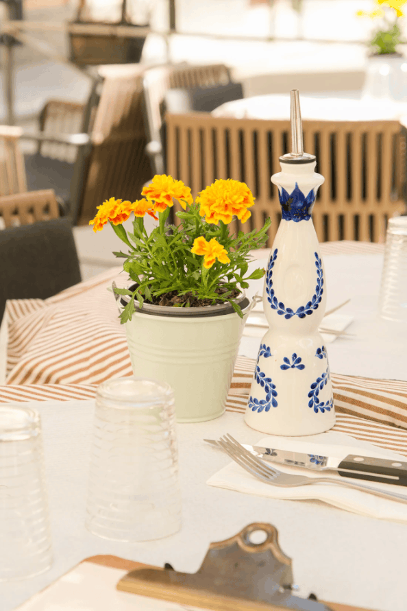 Une table décorée avec un pot de fleurs contenant des fleurs jaunes, un vaporisateur blanc et bleu, des verres en plastique, une fourchette et un couteau, avec un napperon rayé marron et blanc.