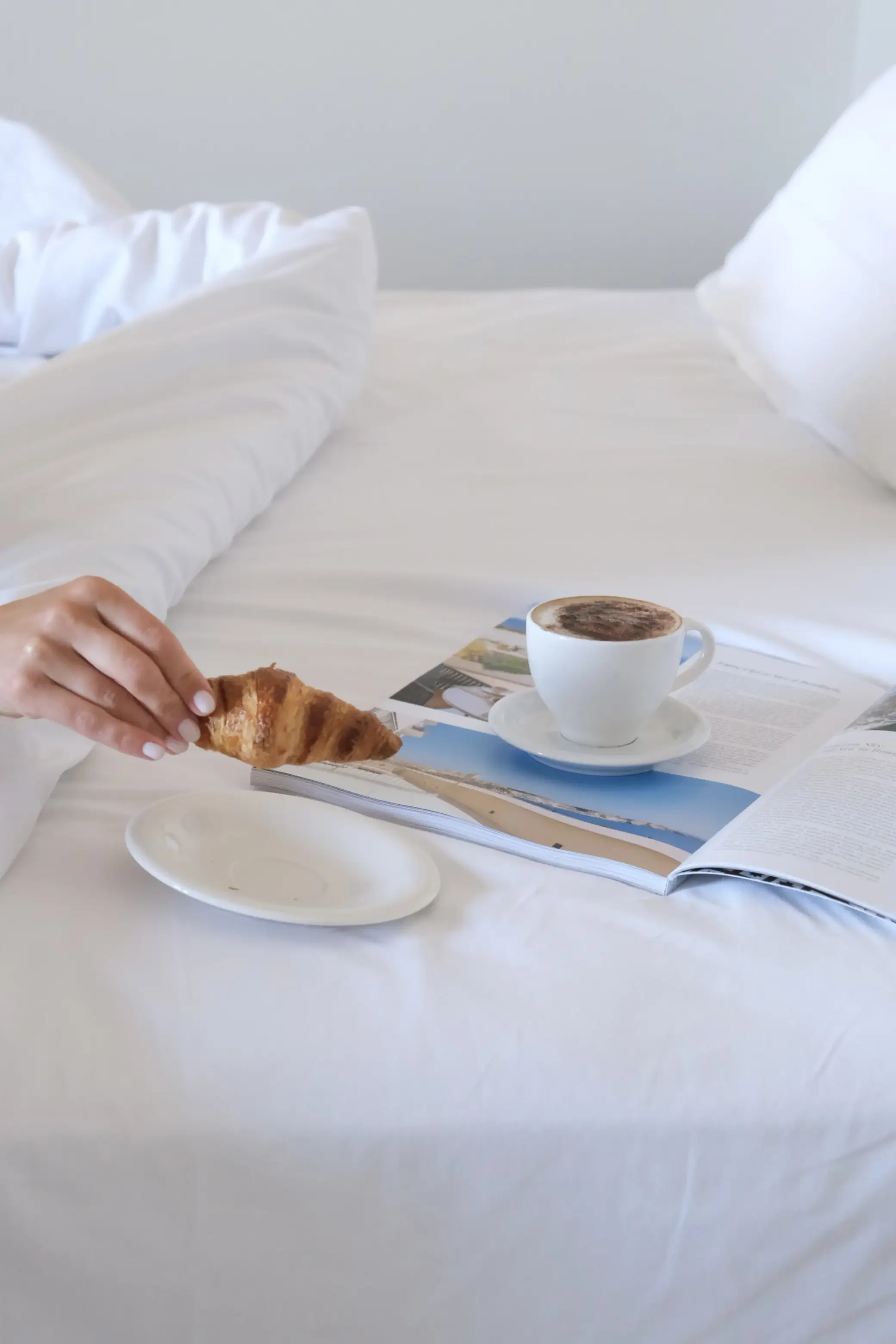Un lit avec des draps blancs, une tasse de café, et un magazine sur la table près du lit.