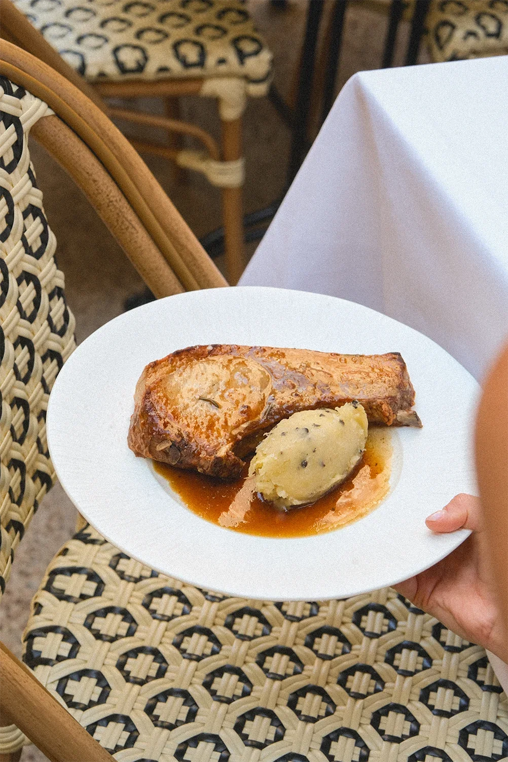 Une assiette avec un morceaux de viande en sauce, une boule de purée et une sauce brune, tenue par une main. En arrière-plan, des chaises en bois et un tissu à motif léopard.